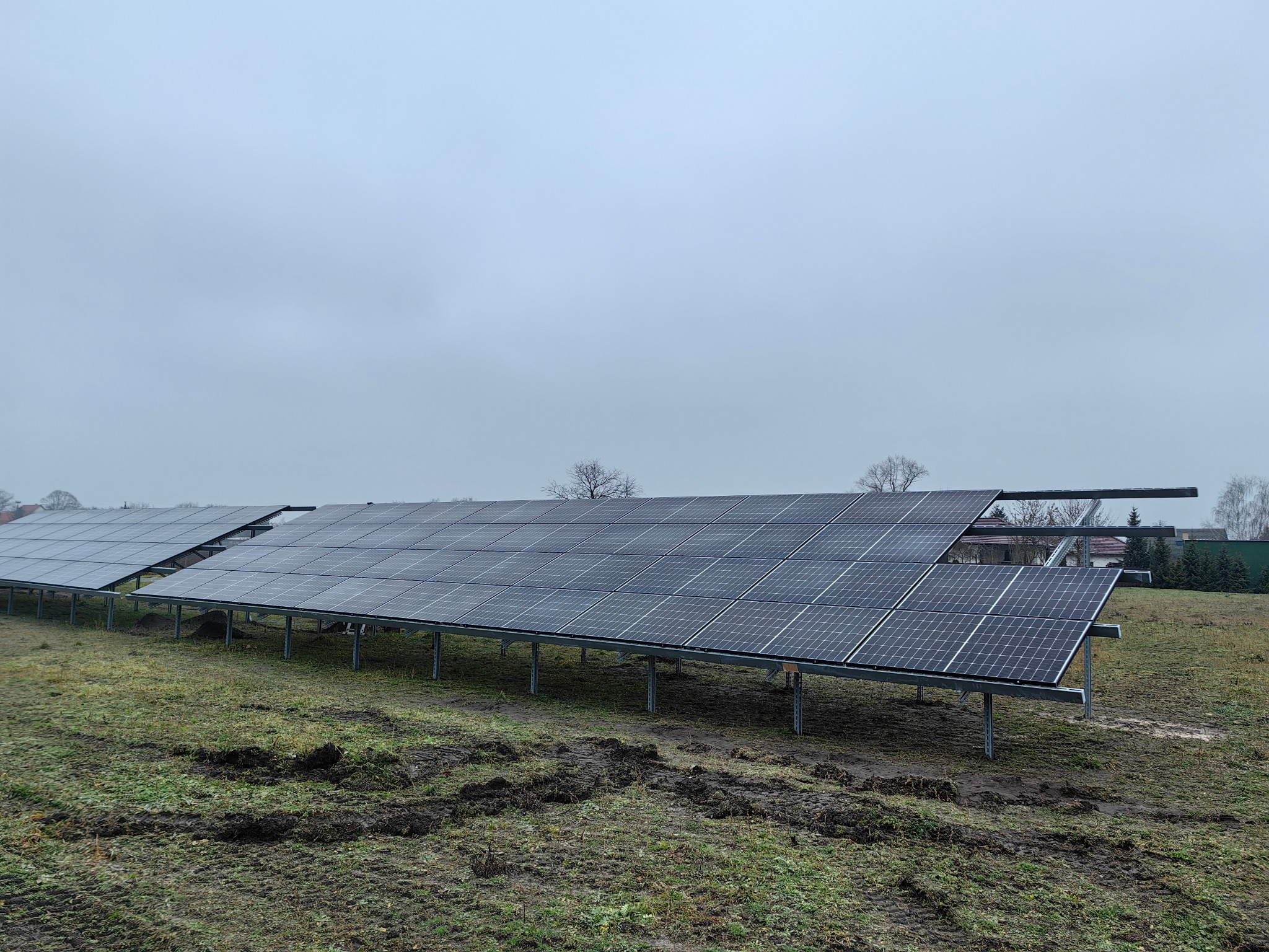 Rząd paneli fotowoltaicznych na metalowej konstrukcji, zainstalowanych na polu w pochmurny dzień. Widoczne ślady opon w błotnistym gruncie.