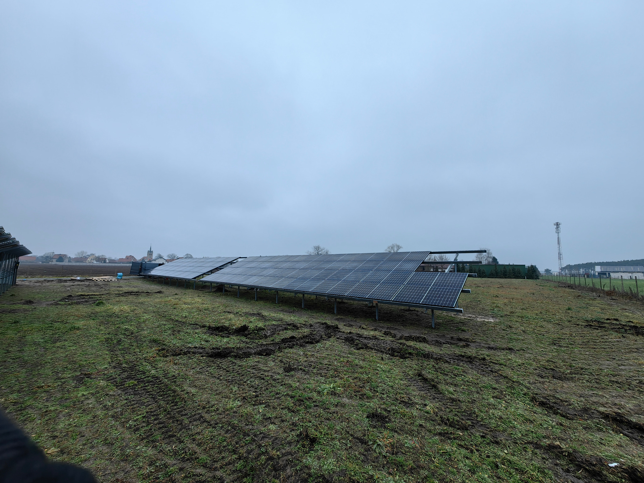 Rząd paneli fotowoltaicznych na metalowej konstrukcji, zainstalowanych na trawiastym terenie z widocznymi śladami opon, pod pochmurnym niebem. W tle zabudowania wiejskie.