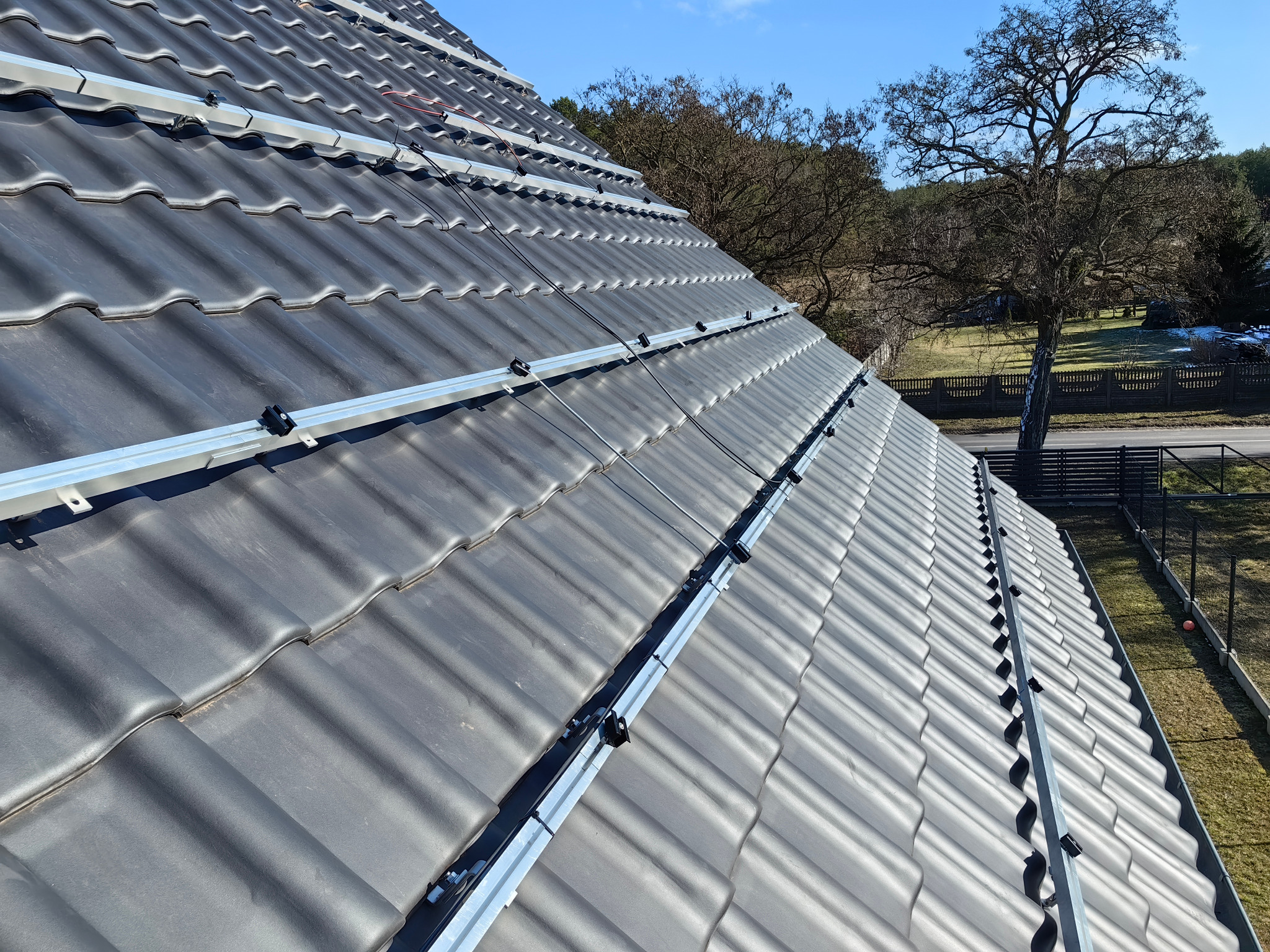 Montaż szyn pod panele fotowoltaiczne na dachu z szarej dachówki ceramicznej. Widoczne elementy mocujące i fragment krajobrazu w tle.