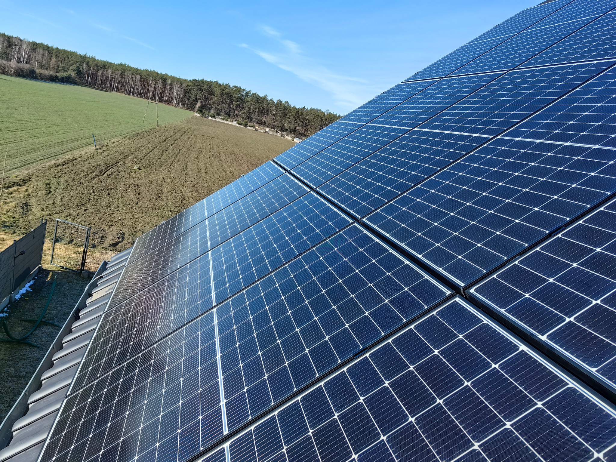 Panel słoneczny na dachu domu, z widokiem na zielone pole i las w tle, sfotografowany w słoneczny dzień. Detaliczna instalacja fotowoltaiczna.