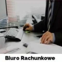 Mężczyzna w garniturze pracuje przy biurku, używając myszy komputerowej i przeglądając dokumenty. Na biurku widoczne są również klawiatura i długopis. W tle żaluzje okienne.