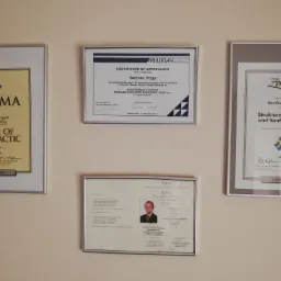 Ściana z czterema oprawionymi dyplomami i certyfikatami, w tym dyplom Master of Chiropractic, certyfikat uczestnictwa w warsztatach Mulligan Concept, dyplom ukończenia kursu masażu terapeutycznego...