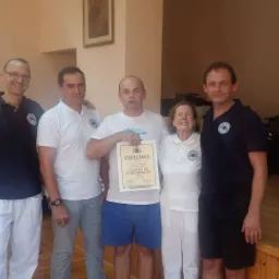 Grupa osób, w tym kobieta i mężczyźni, pozuje z dyplomem Master of Chiropractic w jasnym pomieszczeniu z elementami biurowymi i prezentacyjnymi.