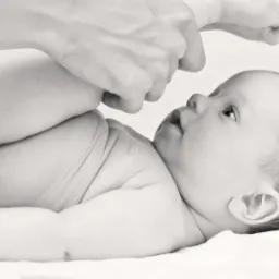 Czarno-białe zdjęcie leżącego na plecach niemowlęcia, którego stopy trzymane są w górze przez dłoń dorosłego, dziecko patrzy w górę.