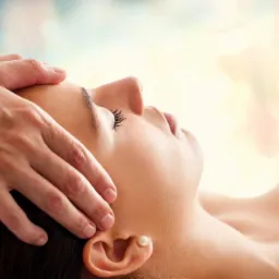 Kobieta z zamkniętymi oczami podczas delikatnego masażu głowy, widoczny fragment perłowego kolczyka w uchu, jasne tło.