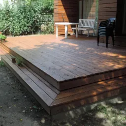 Drewniany taras z dwoma stopniami, na którym stoją białe meble ogrodowe i czarne krzesła. Widoczne rośliny w doniczkach i cień drzew.