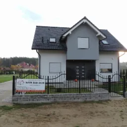 Nowoczesny dom szkieletowy z czarnym dachem i szarą elewacją, otoczony czarnym kutym ogrodzeniem. Widoczny baner firmy budowlanej INTER BUD DOM.