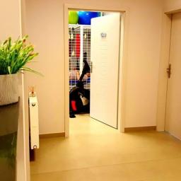 Gabinet Rehabilitacji Leczniczej Medyk Patryk Wydro - Wnętrze gabinetu w Mielcu z widokiem na salę ćwiczeń przez otwarte drzwi, gdzie widoczny jest rower treningowy i regał z piłkami. Na pierwszym planie dekoracyjny kwiat w doniczce i wizytówki firmy.