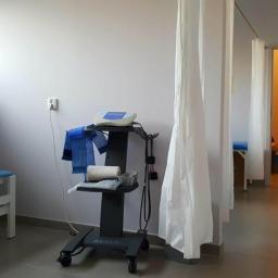 Gabinet Rehabilitacji Leczniczej Medyk Patryk Wydro - Gabinet zabiegowy z parawanami, łóżkiem rehabilitacyjnym, wózkiem z aparaturą Astar oraz sprzętem do terapii światłem w tle.
