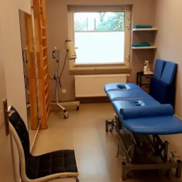 Gabinet fizjoterapeutyczny z niebieskim stołem do masażu, drabinką rehabilitacyjną, lampą terapeutyczną, półkami z ręcznikami i zegarem na ścianie.