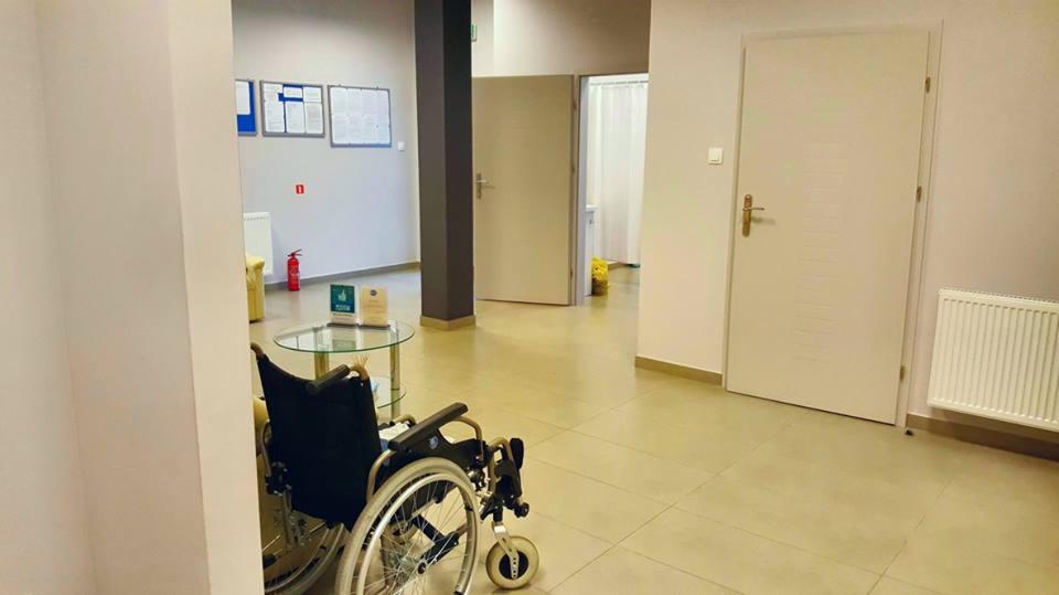 Wnętrze gabinetu rehabilitacyjnego z wózkiem inwalidzkim, stolikiem ze szklanym blatem, tablicami informacyjnymi na ścianie i gaśnicą w tle.