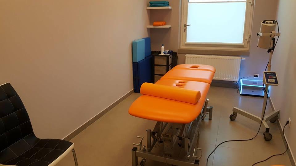 Pomarańczowy stół do masażu w jasnym gabinecie z lampą terapeutyczną i krzesłem w tle.