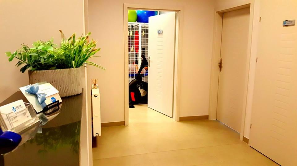 Wnętrze gabinetu w Mielcu z widokiem na salę ćwiczeń przez otwarte drzwi, gdzie widoczny jest rower treningowy i regał z piłkami. Na pierwszym planie dekoracyjny kwiat w doniczce i wizytówki firmy.