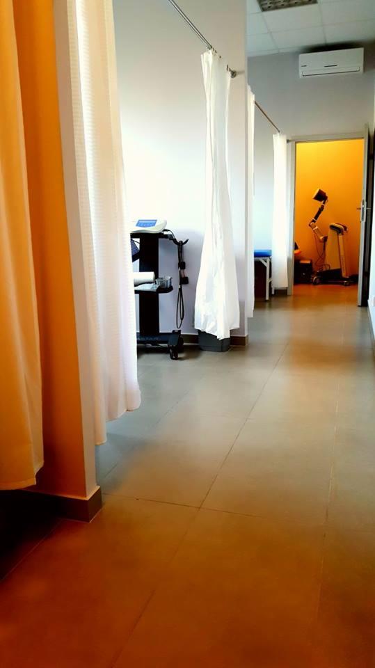 Korytarz w gabinecie z zasłonami oddzielającymi stanowiska, w tle widoczne urządzenia do ćwiczeń rehabilitacyjnych.