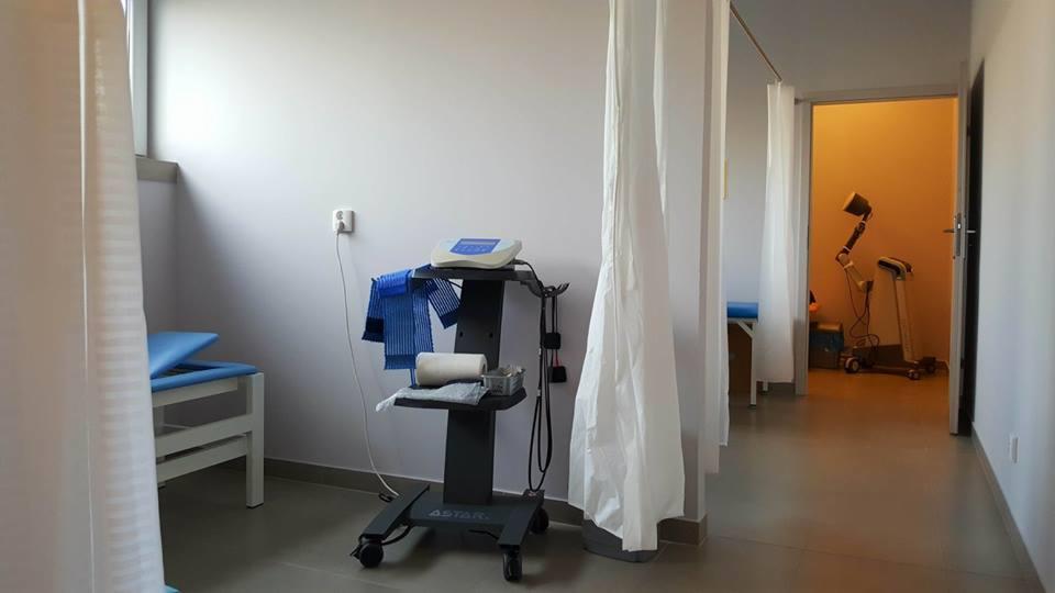 Gabinet zabiegowy z parawanami, łóżkiem rehabilitacyjnym, wózkiem z aparaturą Astar oraz sprzętem do terapii światłem w tle.