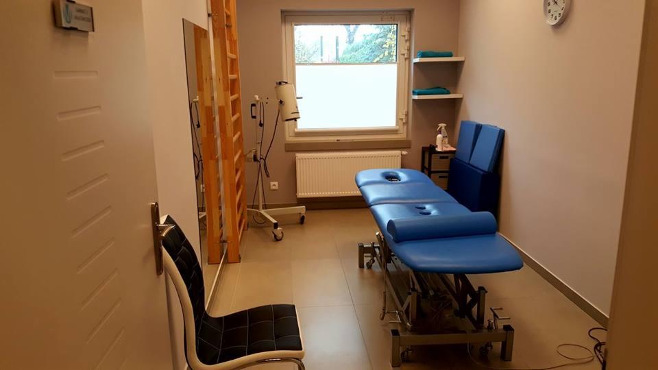 Gabinet fizjoterapeutyczny z niebieskim stołem do masażu, drabinką rehabilitacyjną, lampą terapeutyczną, półkami z ręcznikami i zegarem na ścianie.