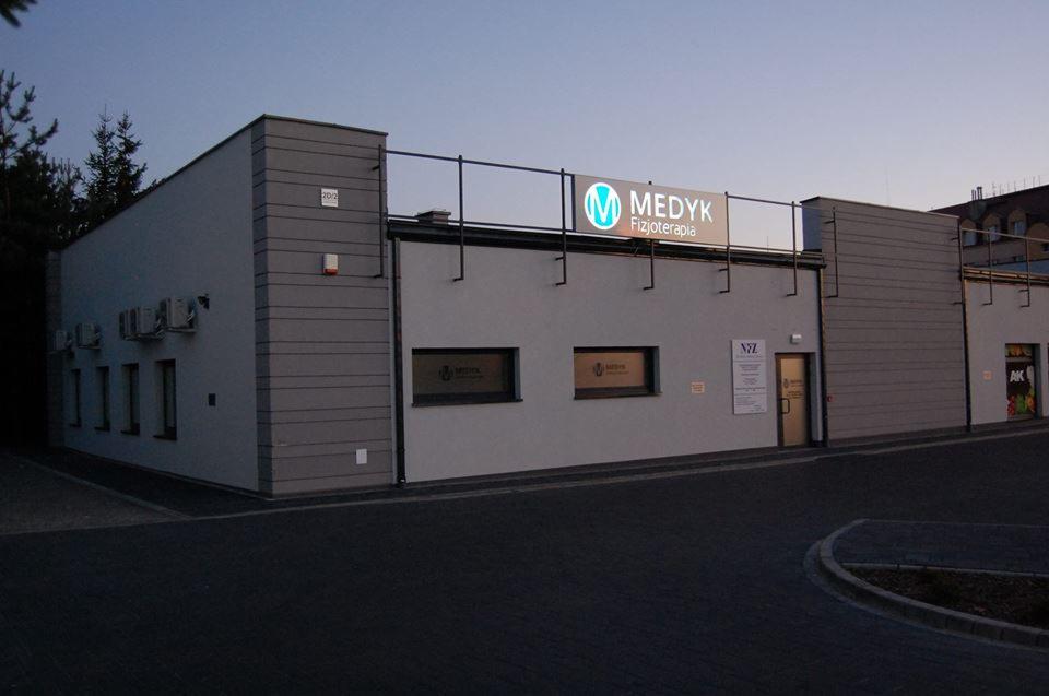 Budynek placówki Medyk Fizjoterapia z podświetlanym szyldem na dachu, widoczne okna z naklejkami informacyjnymi i drzwi wejściowe