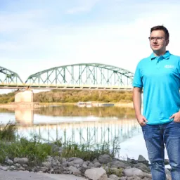 Mężczyzna w okularach i błękitnej koszulce polo z logo firmy na tle rzeki i mostu, stojący na kamienistym brzegu.