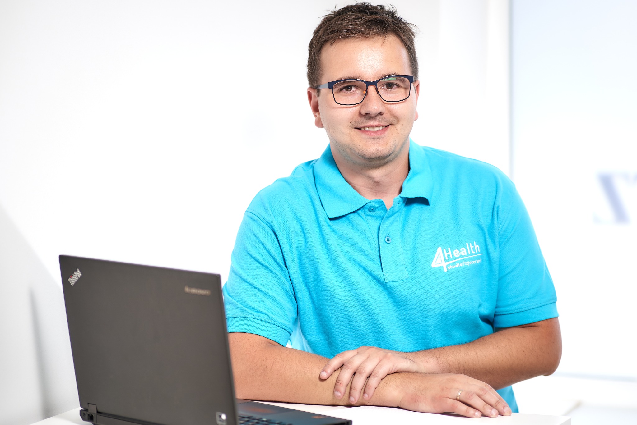 Uśmiechnięty mężczyzna w okularach i błękitnej koszulce polo z logo '4Health studiofizjoterapii' siedzi za biurkiem z laptopem Lenovo ThinkPad.
