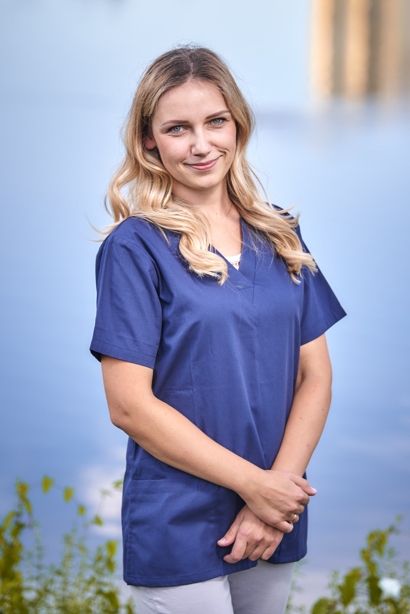 Portret uśmiechniętej blondynki w granatowym uniformie medycznym na tle rozmytego krajobrazu wodnego i zieleni.