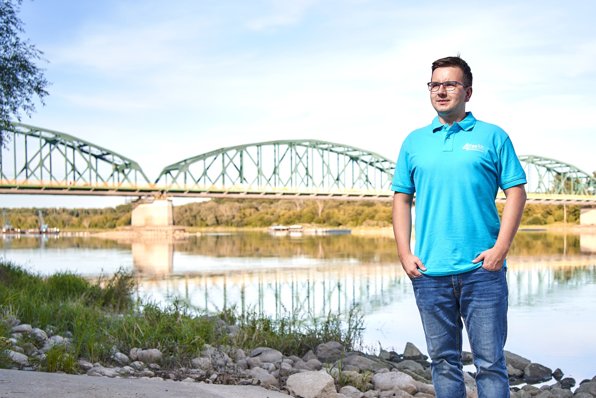 Mężczyzna w okularach i błękitnej koszulce polo z logo firmy na tle rzeki i mostu, stojący na kamienistym brzegu.