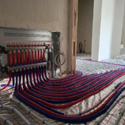 Rozdzielacz z czerwono-niebieskimi rurami w systemie ogrzewania podłogowego, montowany na folii izolacyjnej w surowym wnętrzu budynku.