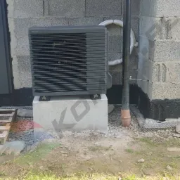 Szara jednostka zewnętrzna urządzenia grzewczego marki Panasonic, zamontowana na betonowym fundamencie przy ścianie z bloczków betonowych, obok drzwi wejściowych i rury spustowej, z drewnianą...