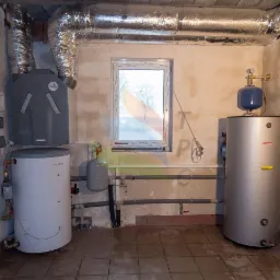 Dwa zbiorniki, instalacja wentylacyjna i okablowanie w pomieszczeniu z oknem. Jeden zbiornik biały, drugi srebrny. Widoczne rury i przewody.