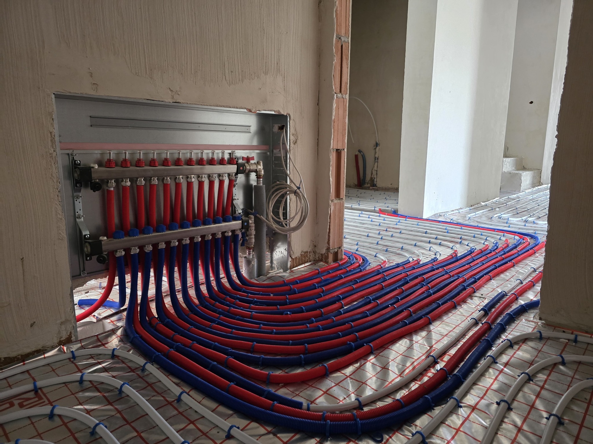 Rozdzielacz z czerwono-niebieskimi rurami w systemie ogrzewania podłogowego, montowany na folii izolacyjnej w surowym wnętrzu budynku.