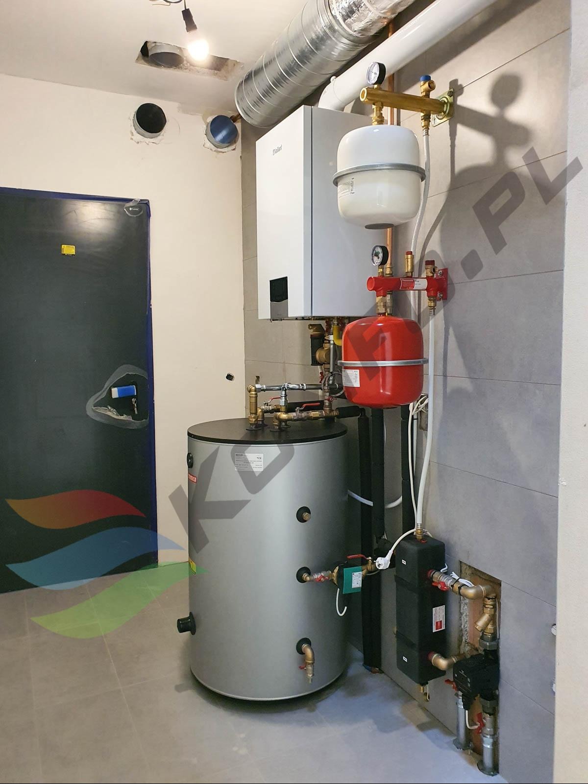 Kompletna instalacja grzewcza z zasobnikiem ciepłej wody, kotłem gazowym i elementami hydraulicznymi zamontowana w pomieszczeniu z szarą podłogą i częściowo wykończonymi ścianami.