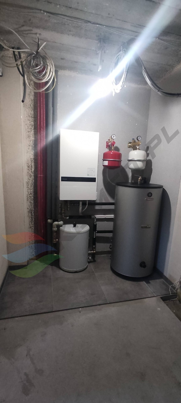 Kotłownia z białym piecem gazowym, srebrnym zbiornikiem, czerwonym i białym naczyniem przeponowym, rurami miedzianymi oraz instalacją elektryczną pod betonowym sufitem.