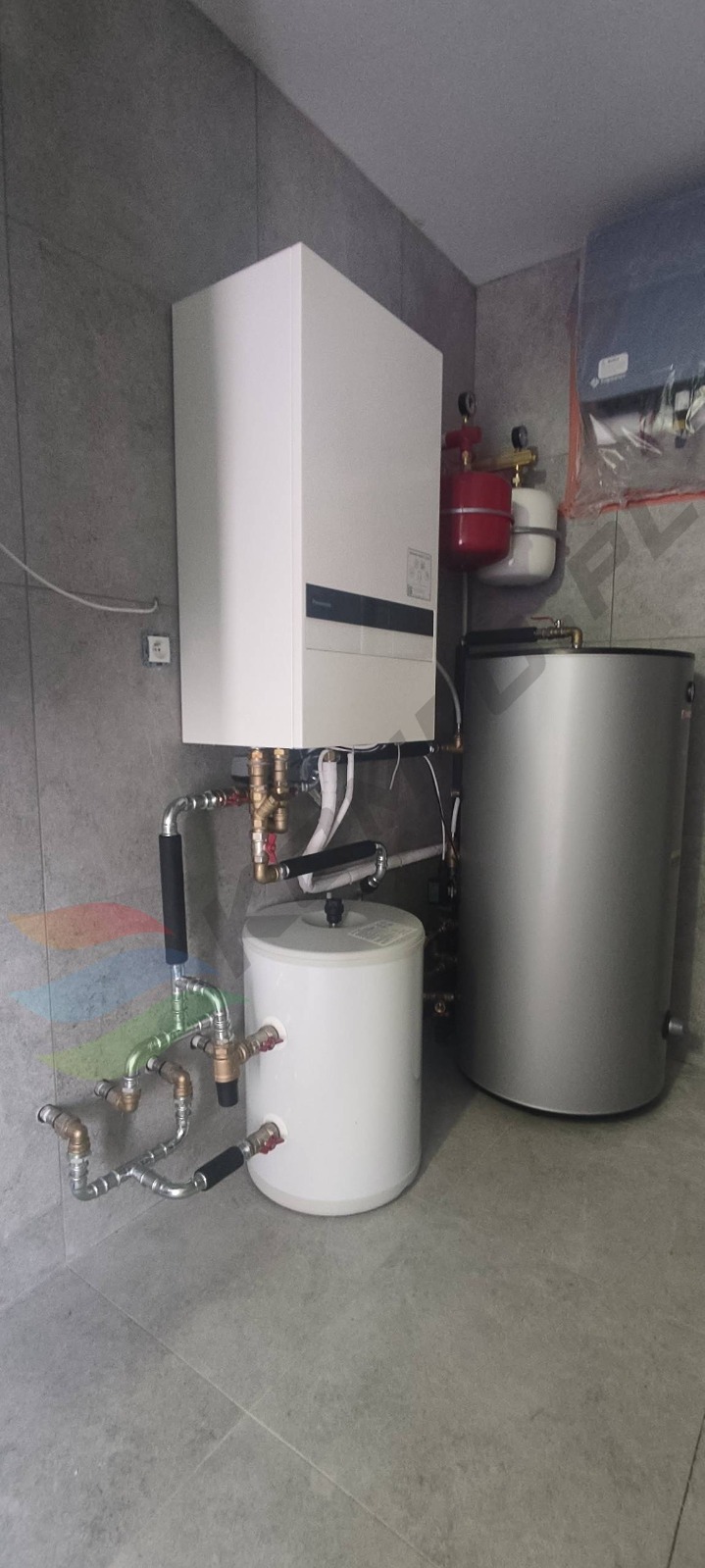 Nowoczesny, kompaktowy system grzewczy z białym kotłem i szarym zasobnikiem na tle szarych płytek w Bielsku-Białej. Widoczne liczne miedziane rury i zawory.