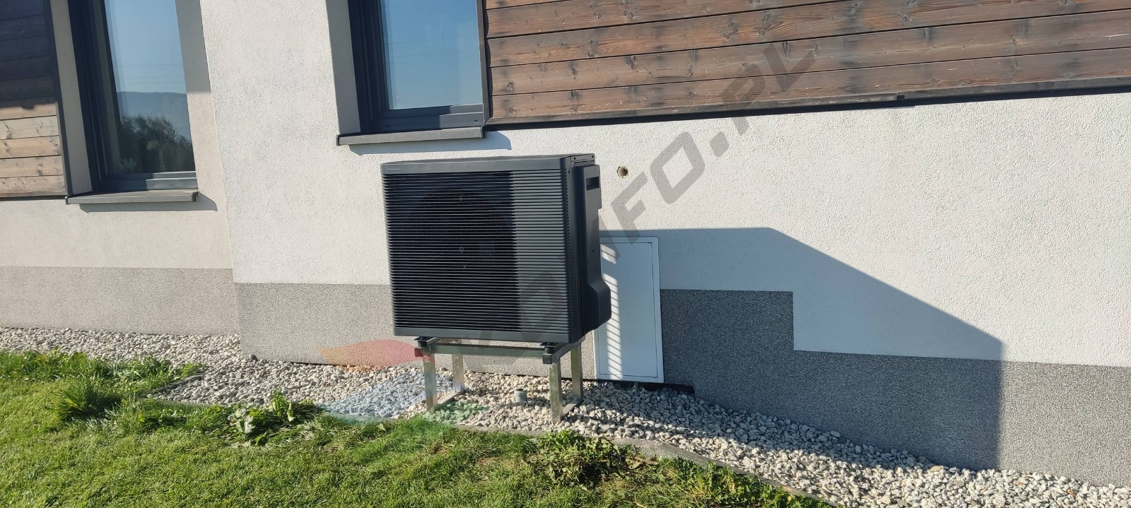 Czarna jednostka zewnętrzna pompy ciepła na metalowej konstrukcji, umieszczona przy ścianie budynku z oknem, na tle trawnika i kamieni ozdobnych.