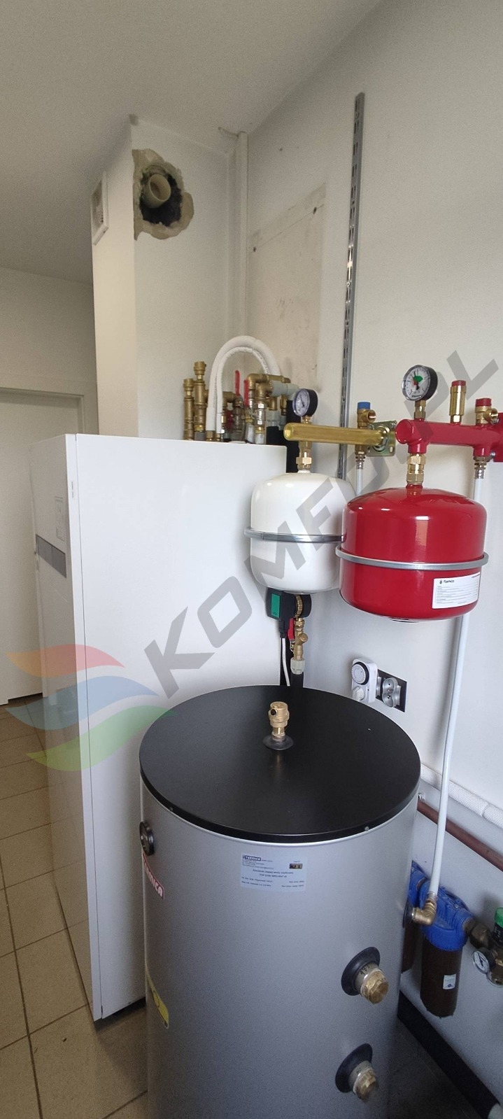 Biała jednostka grzewcza z zbiornikiem akumulacyjnym, czerwonym i białym naczyniem przeponowym, instalacją hydrauliczną z manometrami i zaworami w pomieszczeniu technicznym z uszkodzoną ścianą.