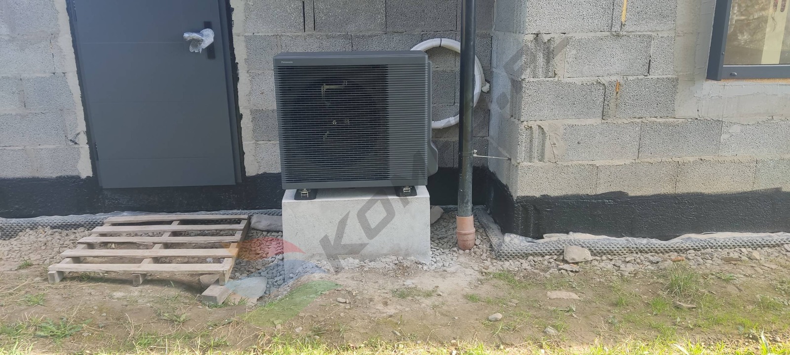 Szara jednostka zewnętrzna urządzenia grzewczego marki Panasonic, zamontowana na betonowym fundamencie przy ścianie z bloczków betonowych, obok drzwi wejściowych i rury spustowej, z drewnianą...