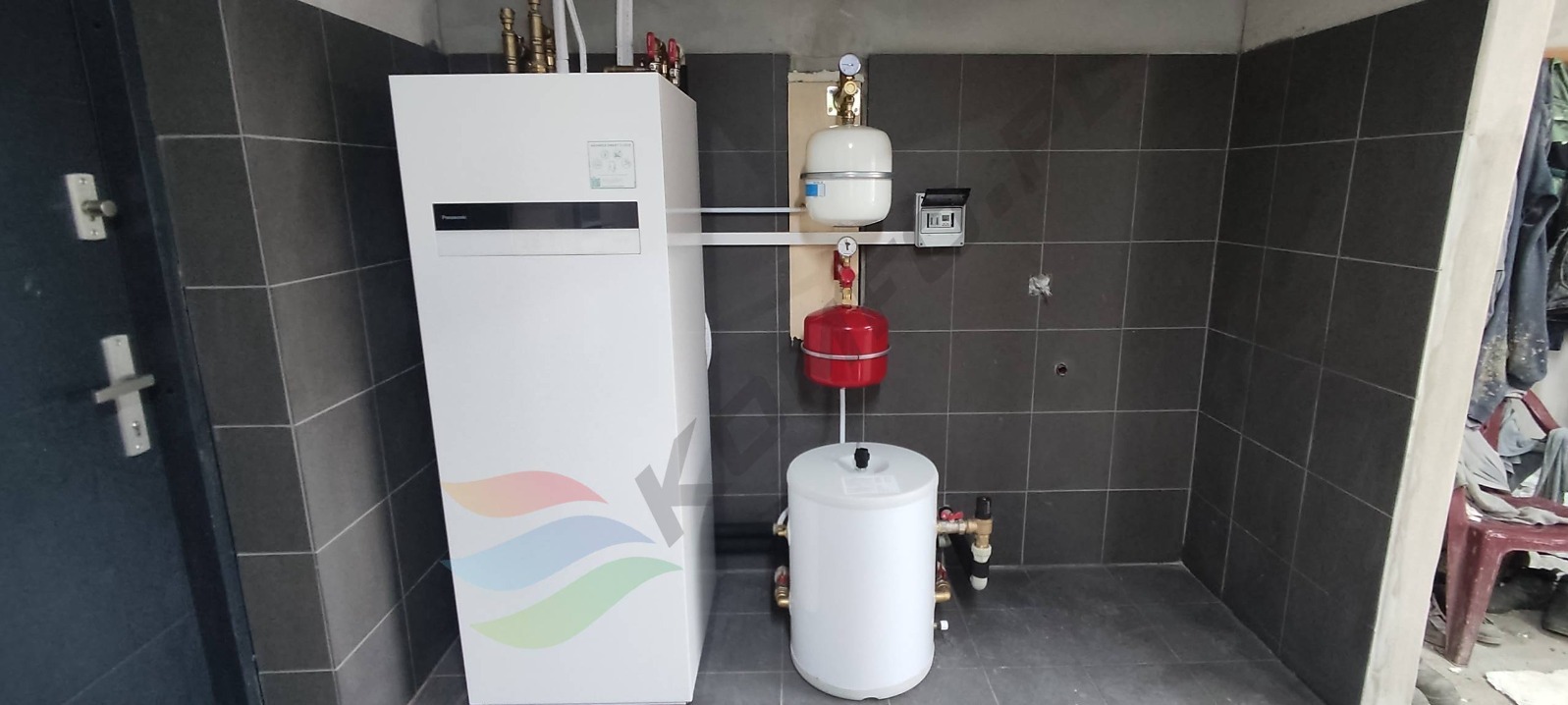 Biała jednostka wewnętrzna z kolorowym logo na tle ciemnych płytek, z widocznymi elementami instalacji hydraulicznymi, takimi jak zbiornik wyrównawczy i naczynie przeponowe.