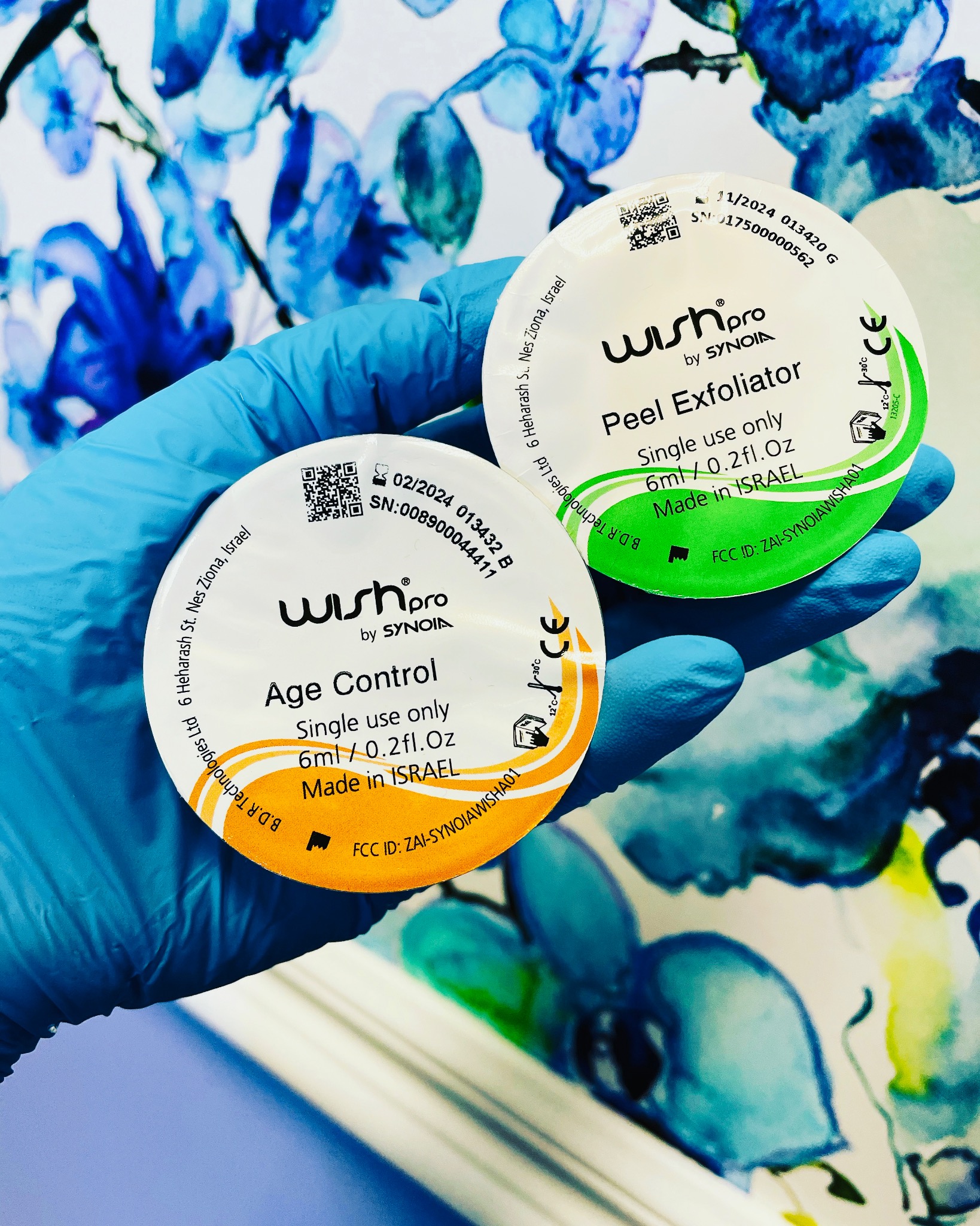Dwa okrągłe opakowania Wishpro by Synoia: zielone 'Peel Exfoliator' i pomarańczowe 'Age Control', trzymane w dłoni w niebieskiej lateksowej rękawiczce na tle białego materiału w niebieskie kwiaty.