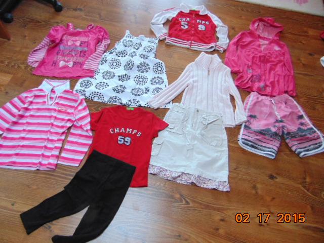 Zestaw używanej odzieży dziecięcej: bluzki, spodnie, spodenki, sukienka i bluzy w kolorach różu, czerwieni i bieli, ułożone na drewnianej podłodze.
