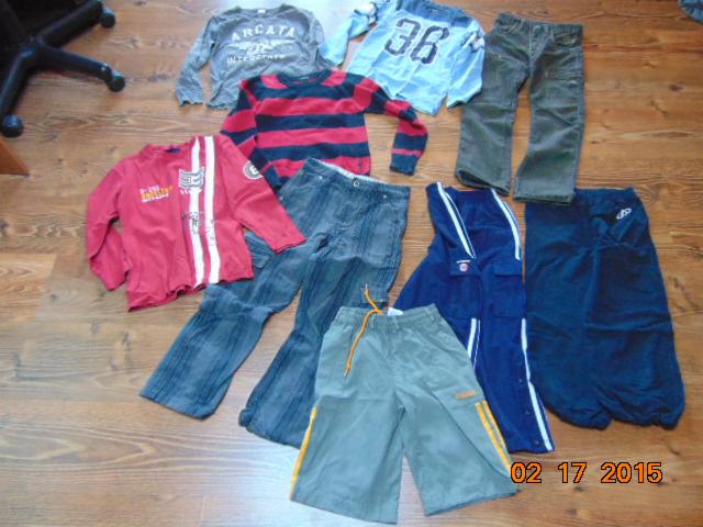 Różnorodny zestaw ubrań dziecięcych, w tym koszulki, spodnie jeansowe i dresowe, ułożonych na drewnianej podłodze. Widoczny fragment krzesła biurowego.