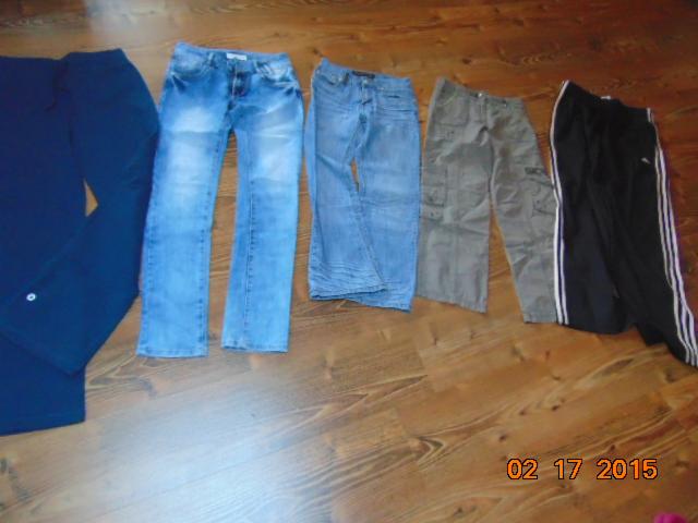 Kolekcja różnorodnych spodni używanych: jeansy, bojówki khaki i dresy Adidas z białymi paskami, ułożone na drewnianej podłodze. Widoczny zapis daty na podłodze.