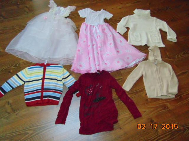 Zestaw ubrań dziecięcych na podłodze: sukienka balowa, sukienka w kwiaty, swetry rozpinane i bluza z kapturem. Różnorodność kolorów i fasonów.