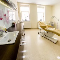 Jasne wnętrze gabinetu kosmetycznego z beżowym fotelem zabiegowym, umywalką z chromowaną baterią i szafkami w kolorze ciemnego drewna, na ścianie grafika z motywem kwiatowym.