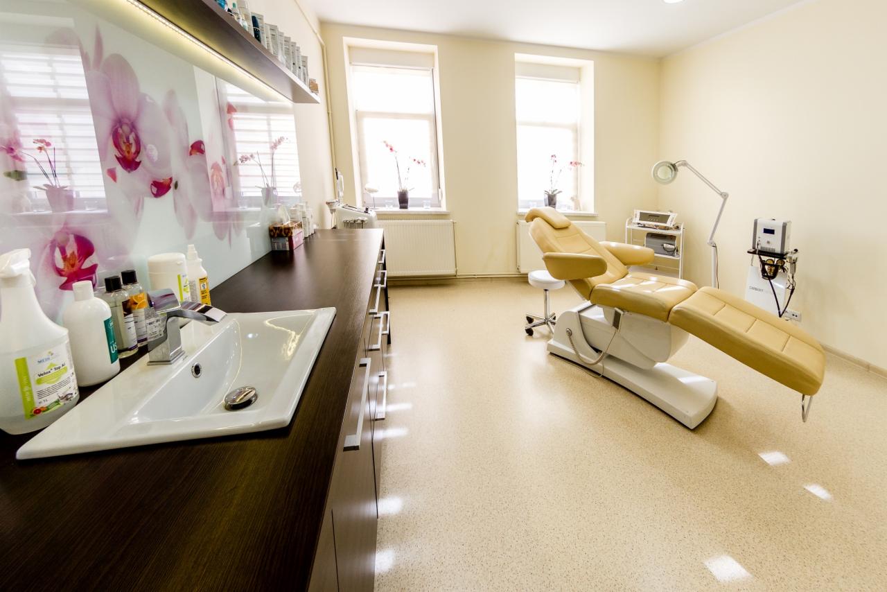 Jasne wnętrze gabinetu kosmetycznego z beżowym fotelem zabiegowym, umywalką z chromowaną baterią i szafkami w kolorze ciemnego drewna, na ścianie grafika z motywem kwiatowym.