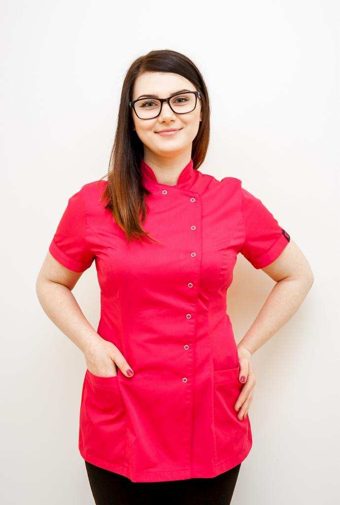 Uśmiechnięta brunetka w okularach, ubrana w czerwoną tunikę kosmetyczną z krótkim rękawem i stójką, pozuje z rękami w kieszeniach na tle białej ściany.