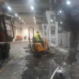 Prace wyburzeniowe w hali w Mogilnie: operator zagęszczarki w żółtej kamizelce wyrównuje podłoże po skuciu betonu, w tle widoczny sprzęt budowlany i pracownicy.
