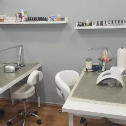 Dwa stanowiska do manicure z lampami UV, frezarką i paletą kolorów lakierów na tle szarych ścian i półek z lakierami w salonie kosmetycznym.