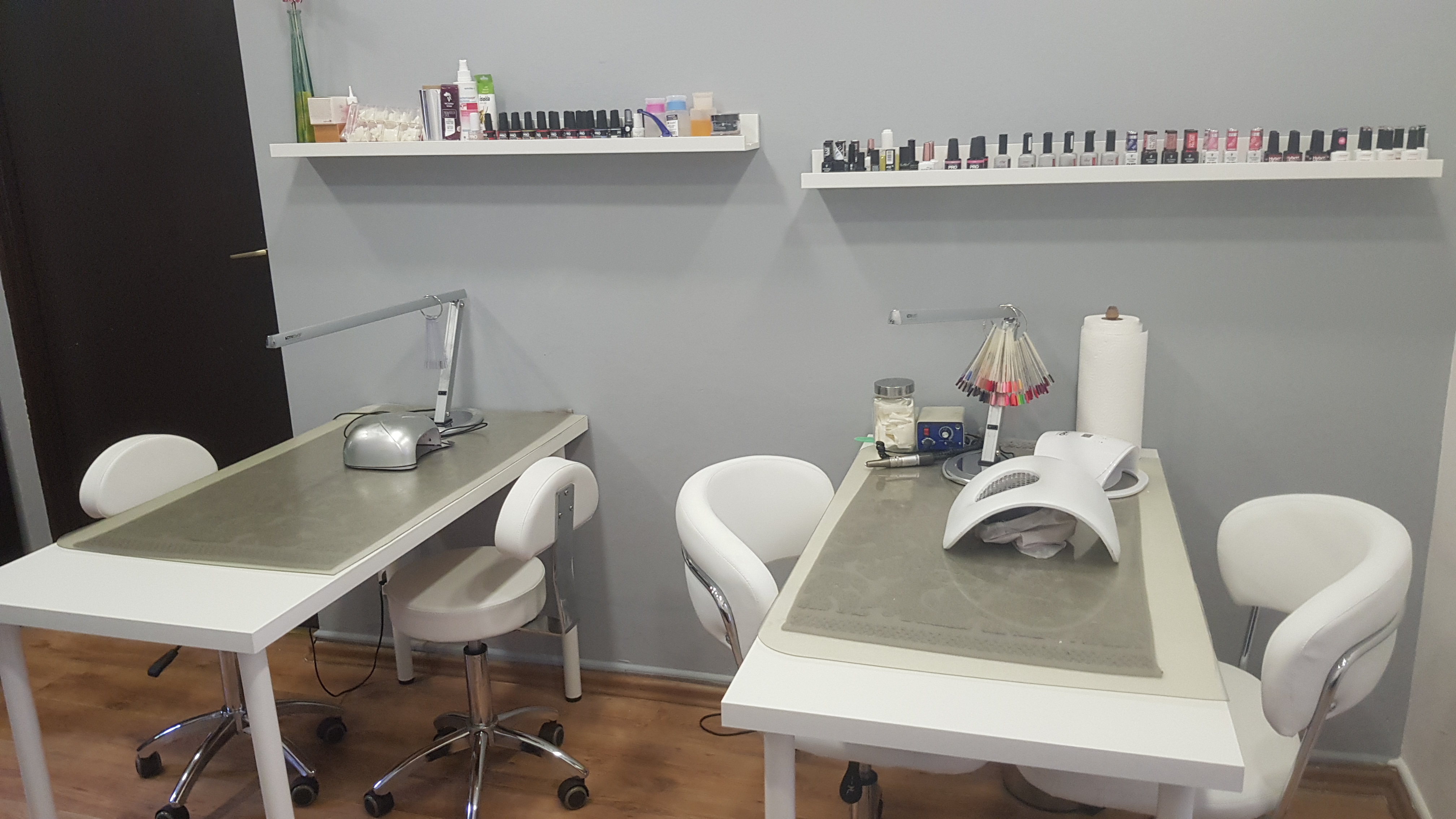 Dwa stanowiska do manicure z lampami UV, frezarką i paletą kolorów lakierów na tle szarych ścian i półek z lakierami w salonie kosmetycznym.