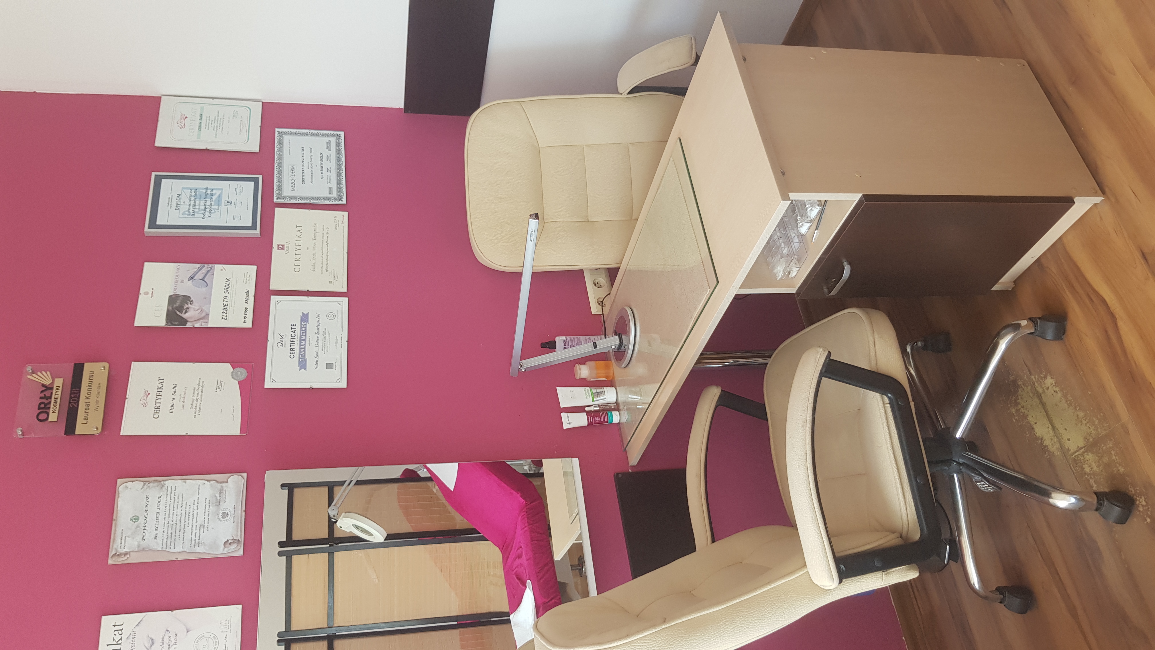 Stanowisko pracy w salonie: biurko z lampą, krzesło obrotowe, na ścianie liczne certyfikaty i dyplomy w ramkach, kosmetyki na blacie.