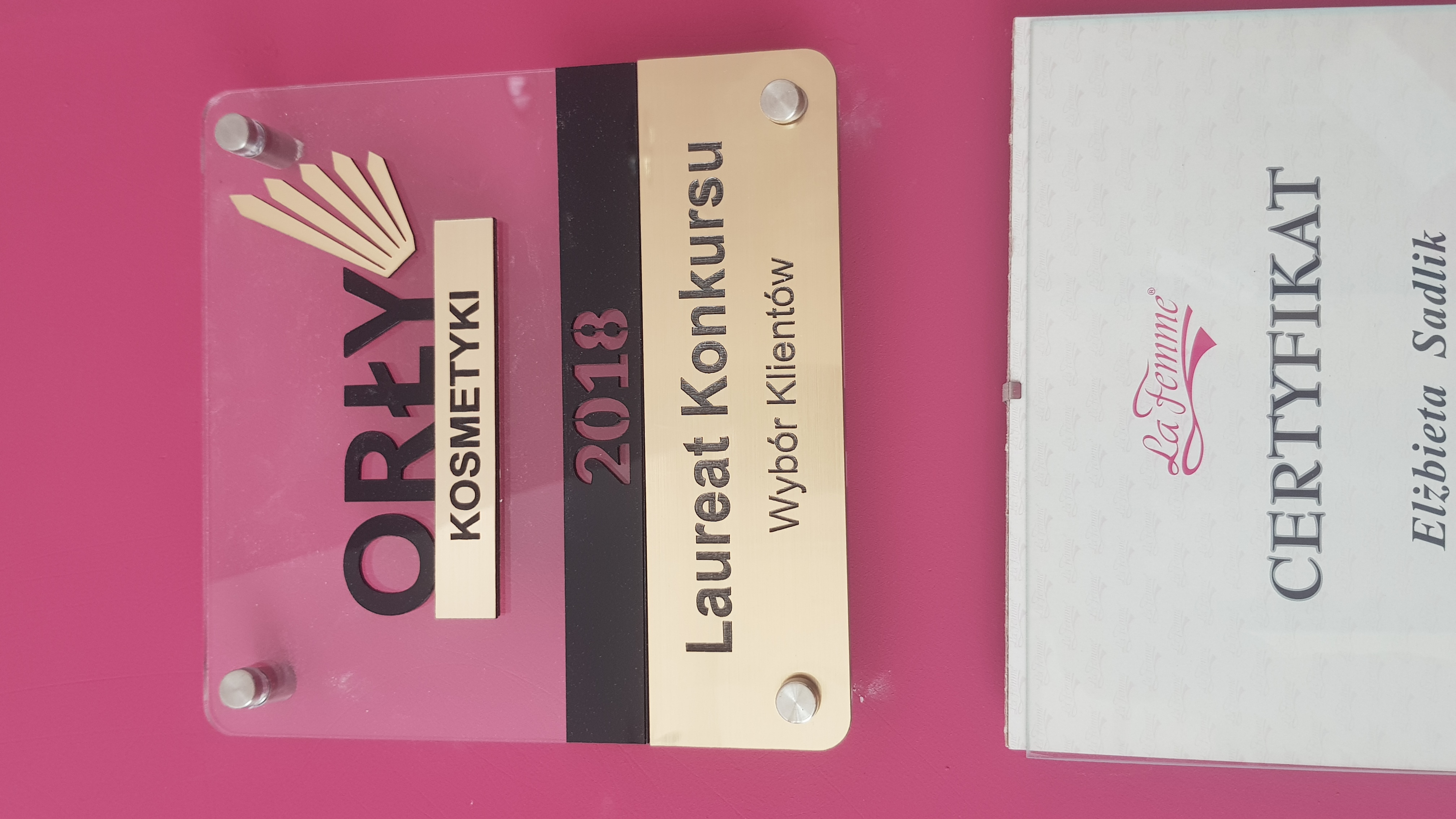Tablica z logo 'Orły Kosmetyki 2018 Laureat Konkursu Wybór Klientów' oraz certyfikat 'La Femme' Elżbieta Sadlik na różowym tle.
