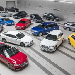 Różnokolorowa flota samochodów marki Audi, w tym kabriolety, sedany, kombi i SUV-y, zaparkowana w przestronnym garażu oświetlonym światłami sufitowymi.